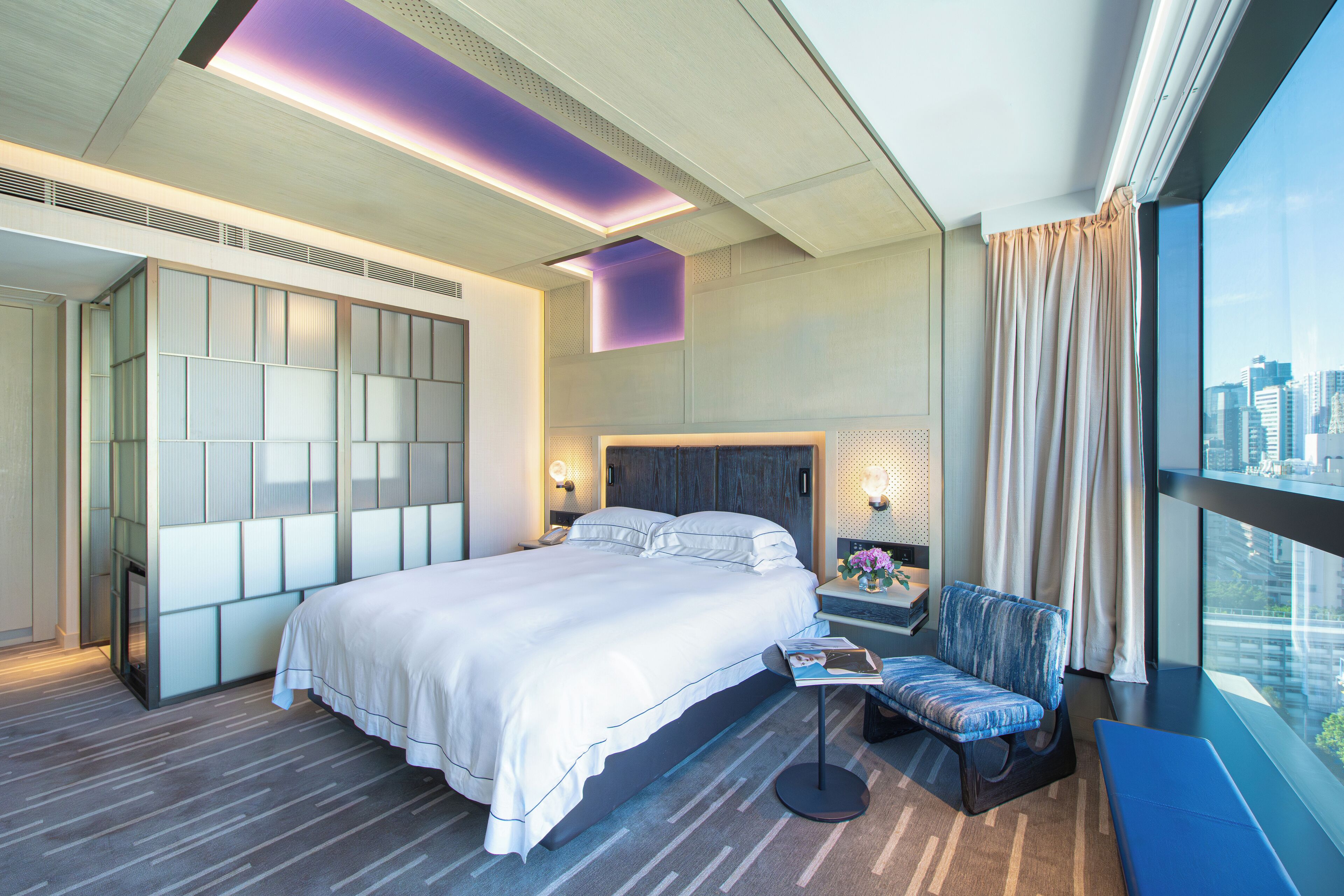 A modern hotel room with a large bed, a seating area with a chair, a nightstand, and a view of the city through large windows.
