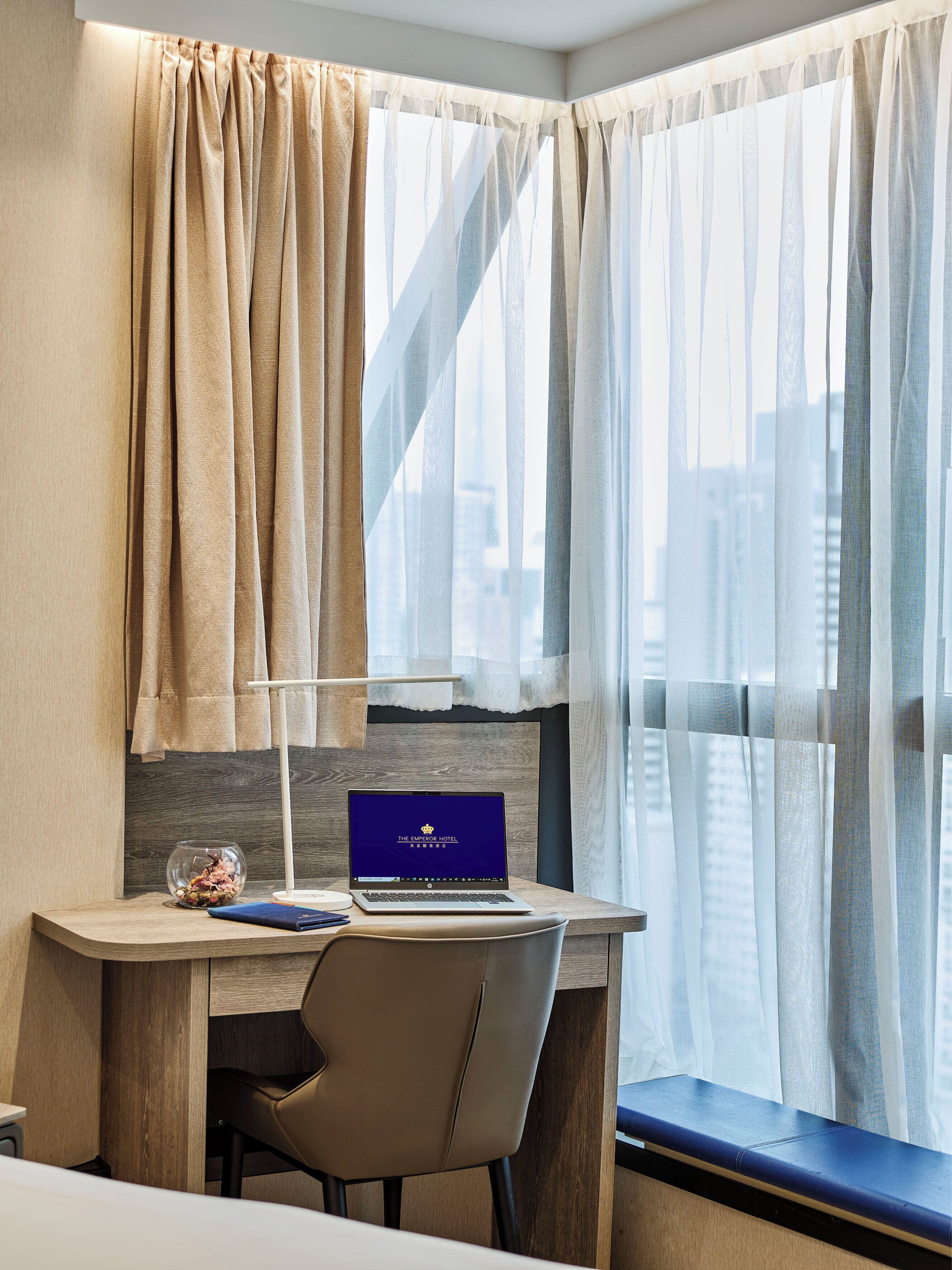 Une chambre d’hôtel équipée d’un bureau, d’un ordinateur portable et offrant une vue sur la ville à travers de fines rideaux.