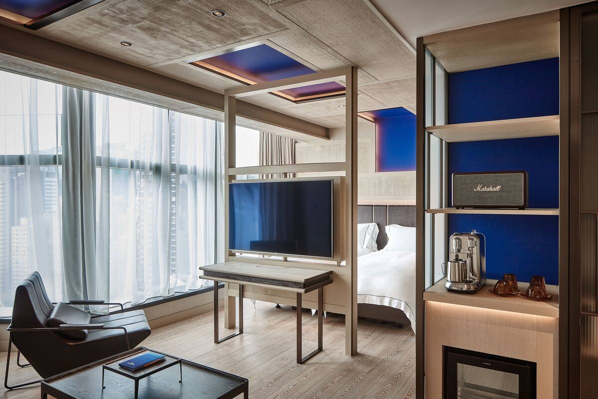 A modern hotel room with a large window, a flat-screen TV, a bed, a coffee machine, and a minibar.