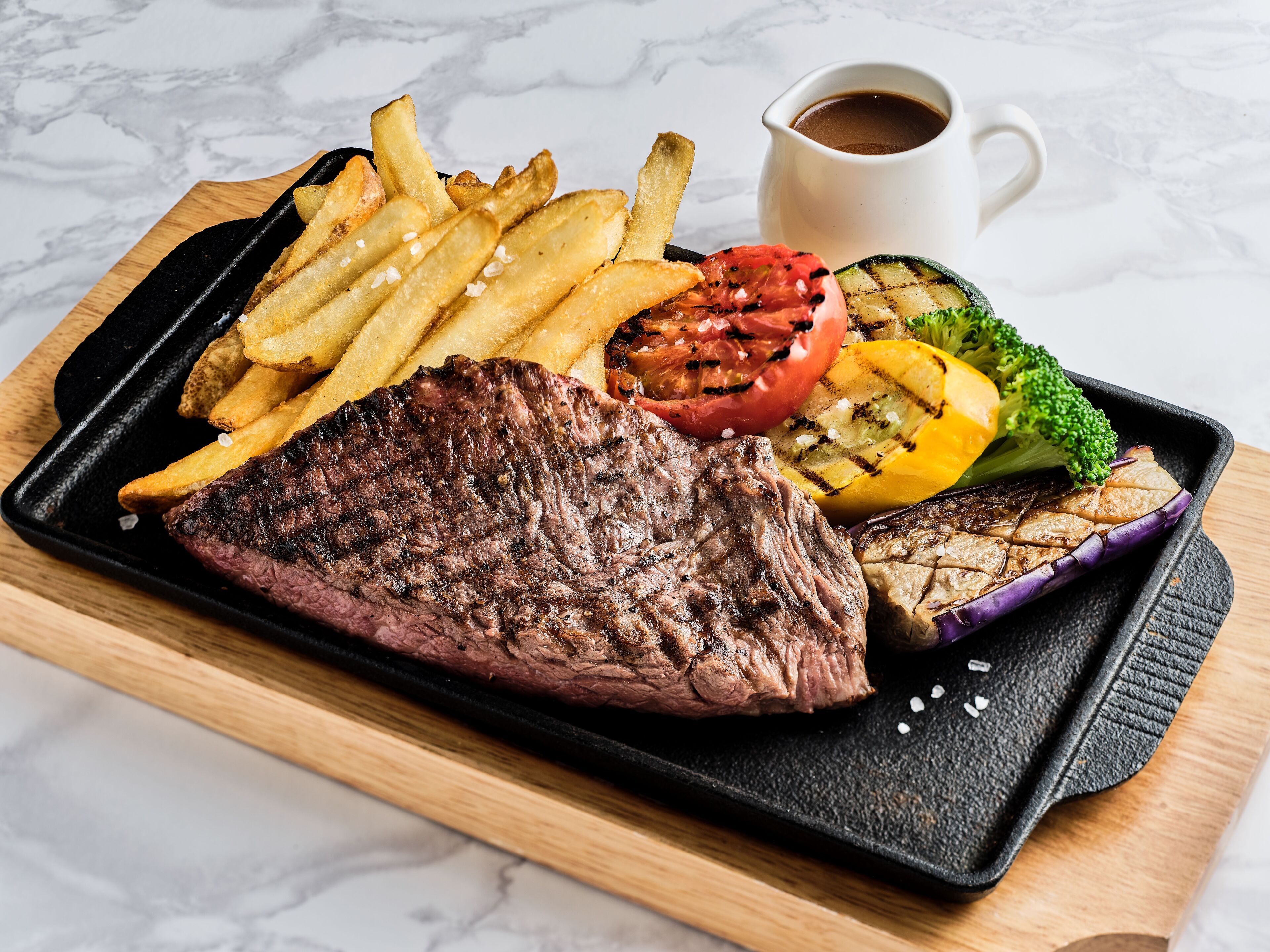 Un bistec a la parrilla, verduras asadas y papas fritas sobre una bandeja negra.