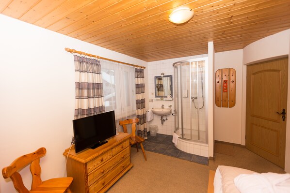 A bedroom with a wooden ceiling, a television on a dresser, a shower, and a wooden door. - DEVA Hotel Kaiserblick (Reit im Winkl)