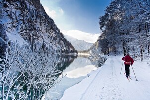 Paisaje nevado con una persona esquiando por un sendero junto a un lago tranquilo, rodeado de árboles cargados de nieve y montañas.
