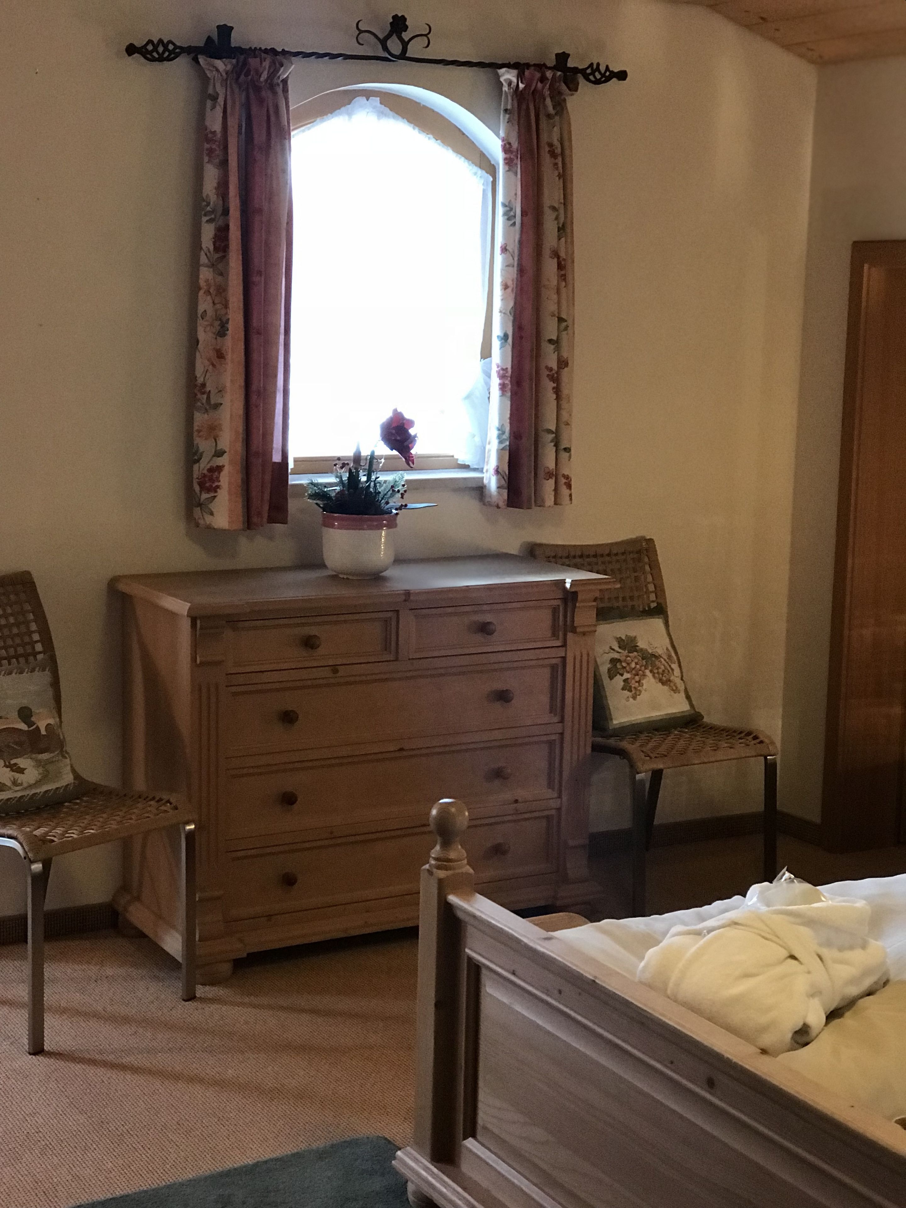 A bedroom with a wooden dresser, two chairs, a bed, and a window with floral curtains.