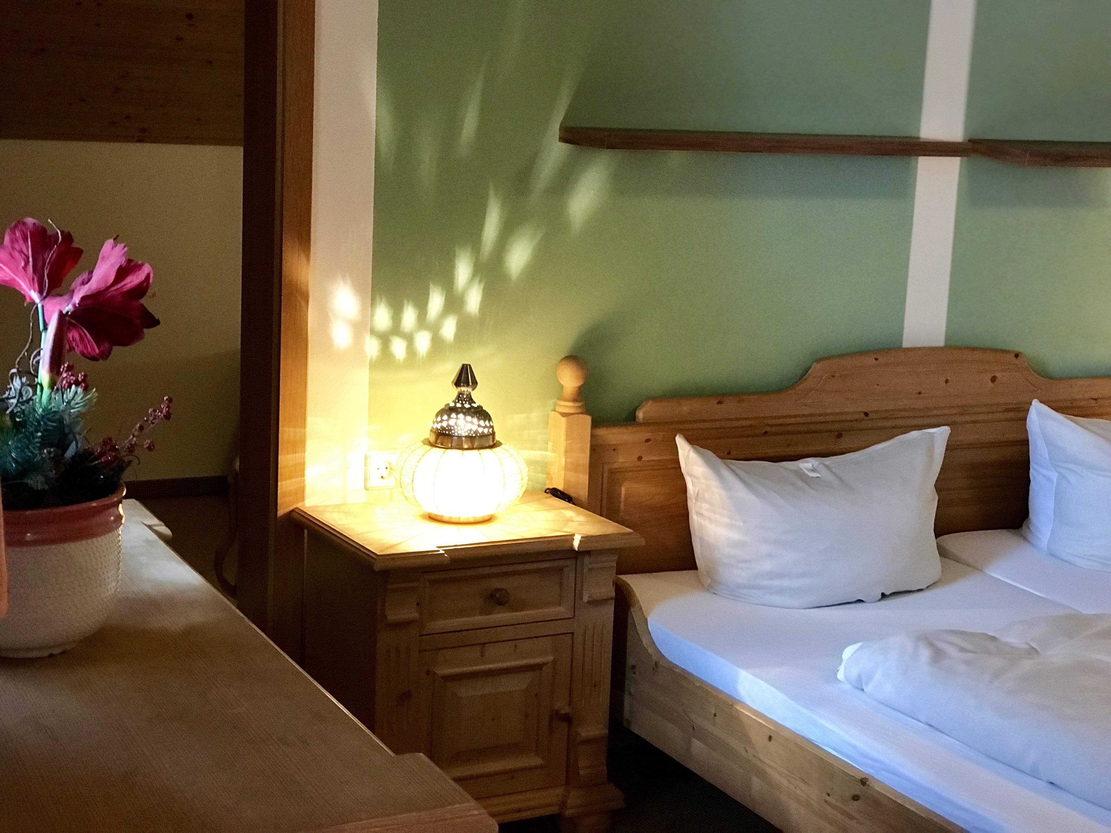 A wooden bed with white bedding, a nightstand with a lamp, and a potted plant with pink flowers.