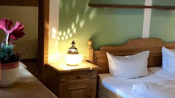 Una cama de madera con ropa de cama blanca, una mesita de noche con una lámpara y una planta en maceta con flores rosadas.