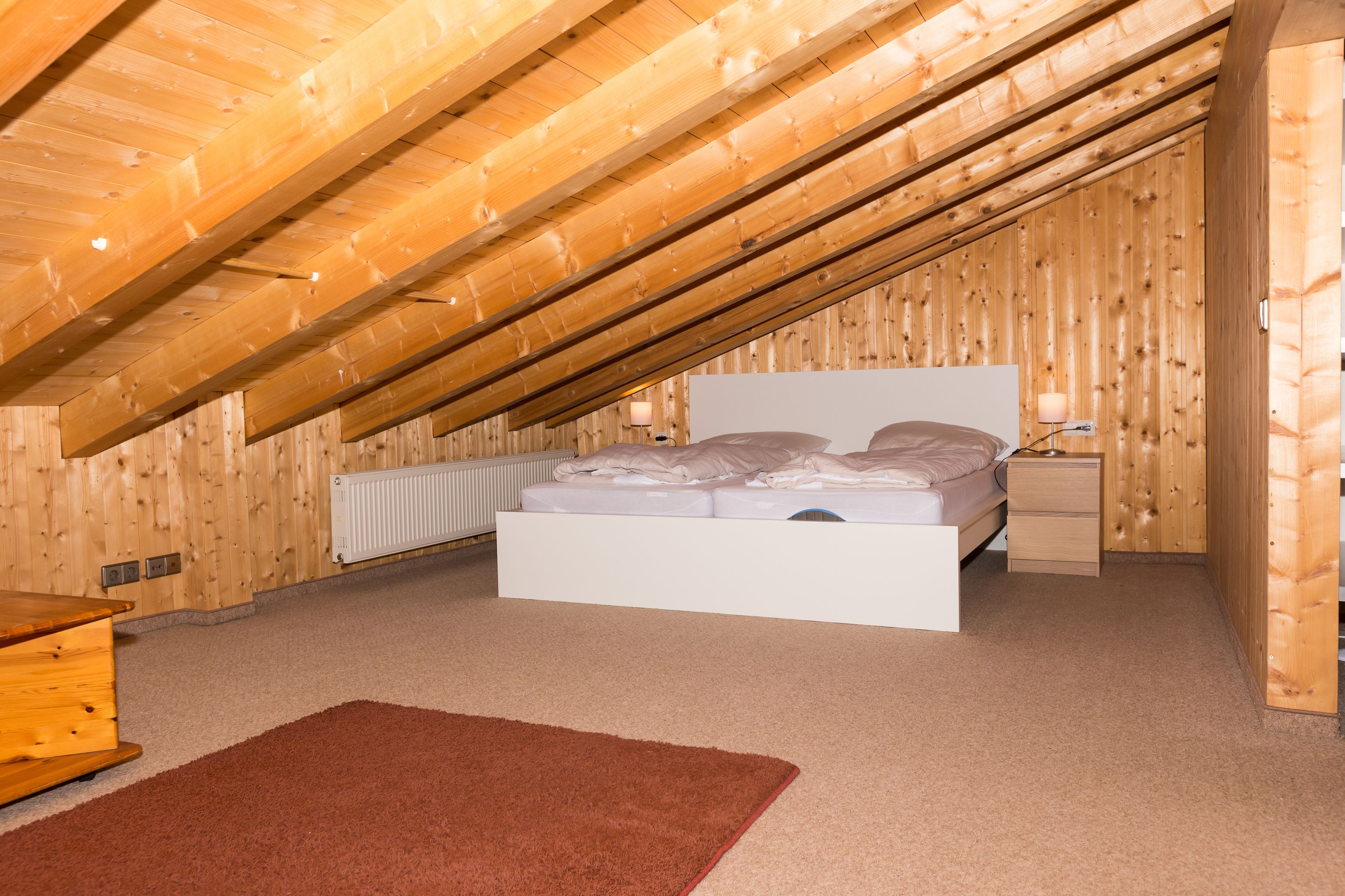 A bedroom with a sloped ceiling, a bed, a bedside table, a lamp, and a radiator.