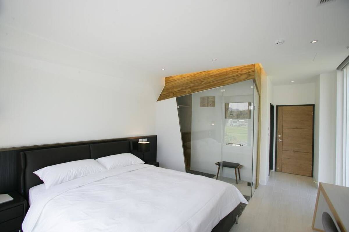 A modern bedroom with a large bed, a nightstand, a wooden ceiling feature, and a glass door leading to a balcony.