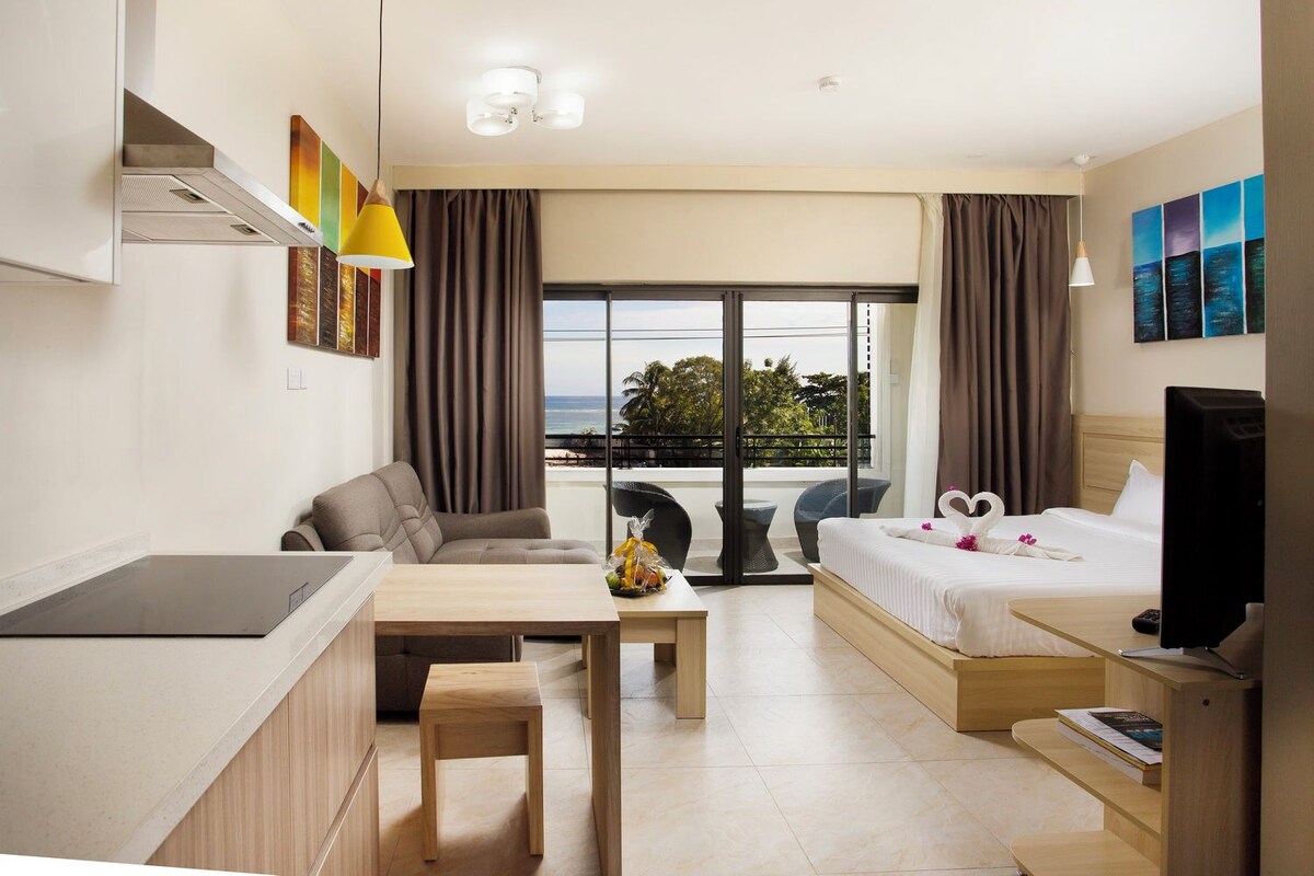Habitación de hotel con kitchenette, sofá, cama, televisión y balcón con vista al océano.