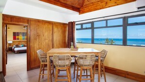Uma sala de jantar com mesa de madeira e cadeiras de vime, vista para o oceano através de grandes janelas e teto de madeira.