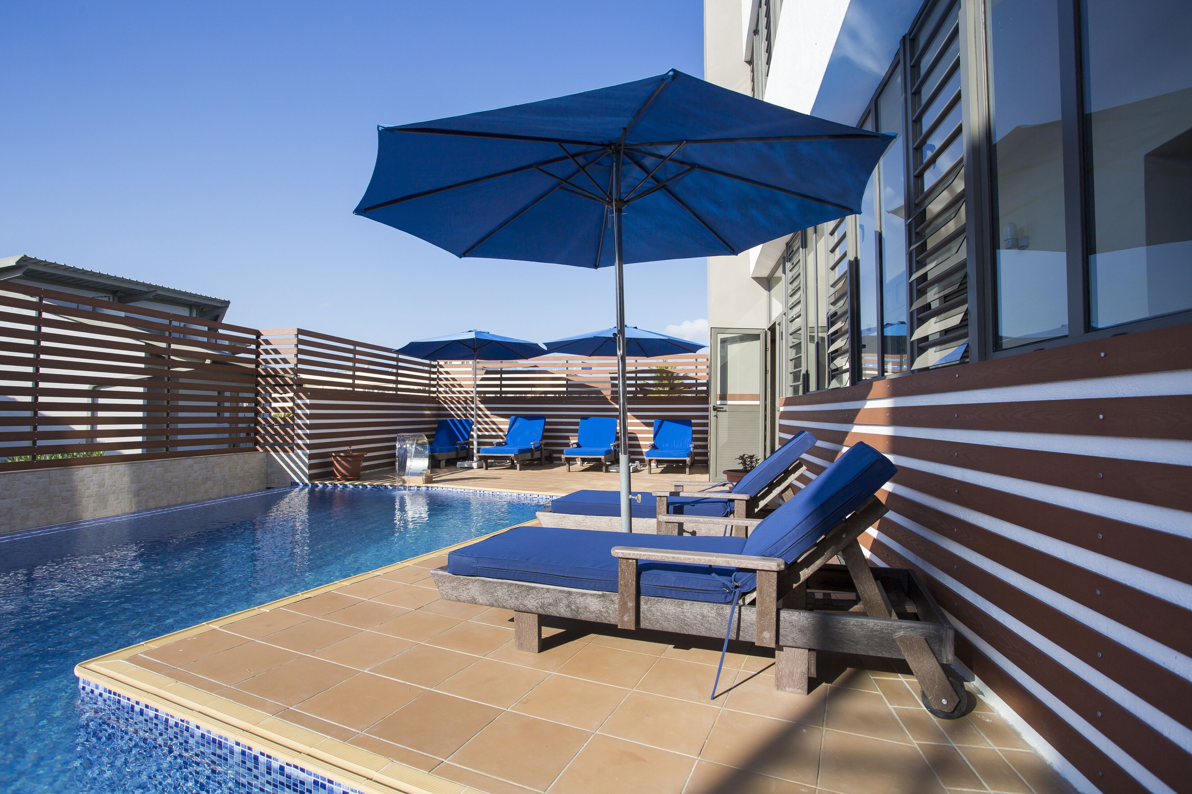 A rooftop pool area with blue umbrellas, lounge chairs, and a clear blue sky.