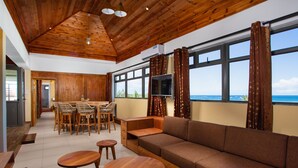 Uma sala de estar espaçosa com teto de madeira, um sofá grande, duas mesas redondas de madeira e um conjunto de jantar com cadeiras de vime.