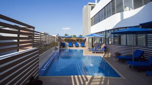 Uma área de piscina com cadeiras de descanso azuis e guarda-sóis, um cercado de madeira e um edifício moderno com grandes janelas.