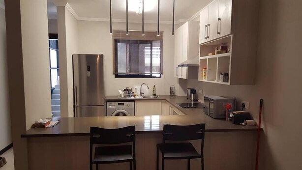 A modern kitchen with a stainless steel refrigerator, a built-in washing machine, a sink, and a microwave. There are two black chairs at the kitchen island.