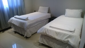 Two single beds with white linens, a nightstand, and a window with curtains. - Lifestyle Apartments (Gaborone)
