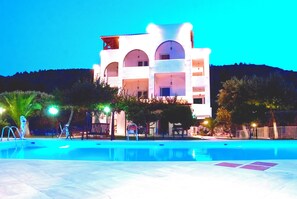 Un hotel con piscina, mobiliario exterior y montañas al fondo.