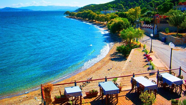 Vista costera con playa, muelle de madera con mesas y sillas, y montaña al fondo.