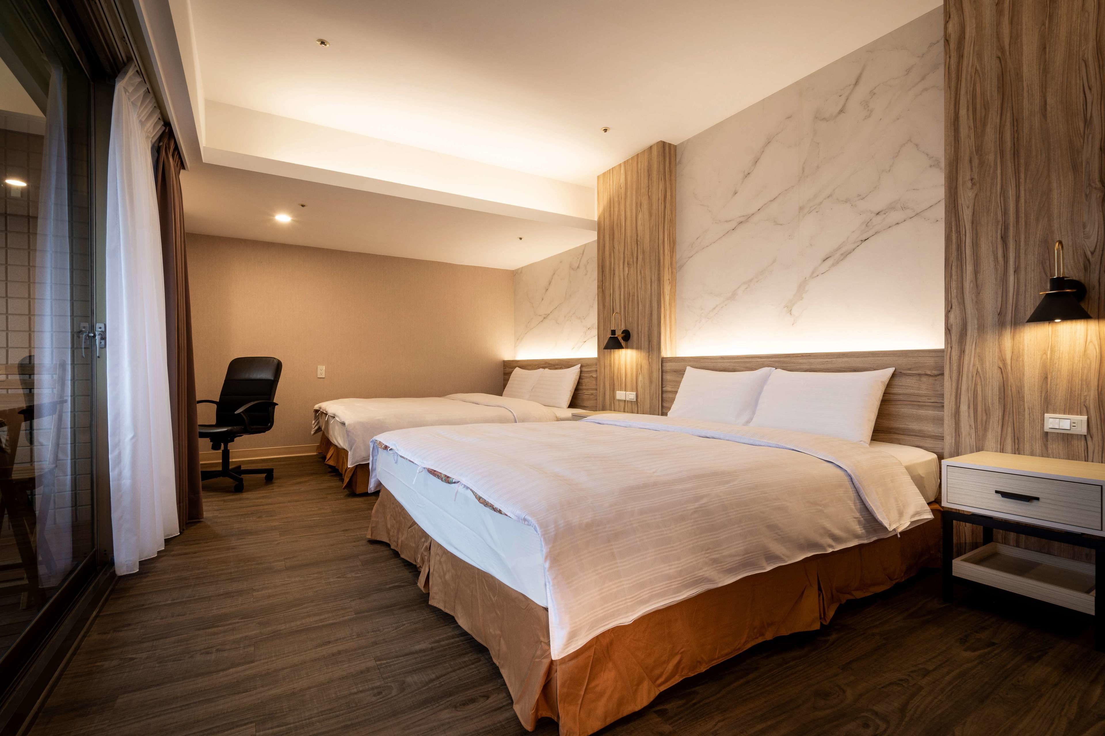 A hotel room with two beds, a wooden floor, a marble wall, and a desk with a lamp.