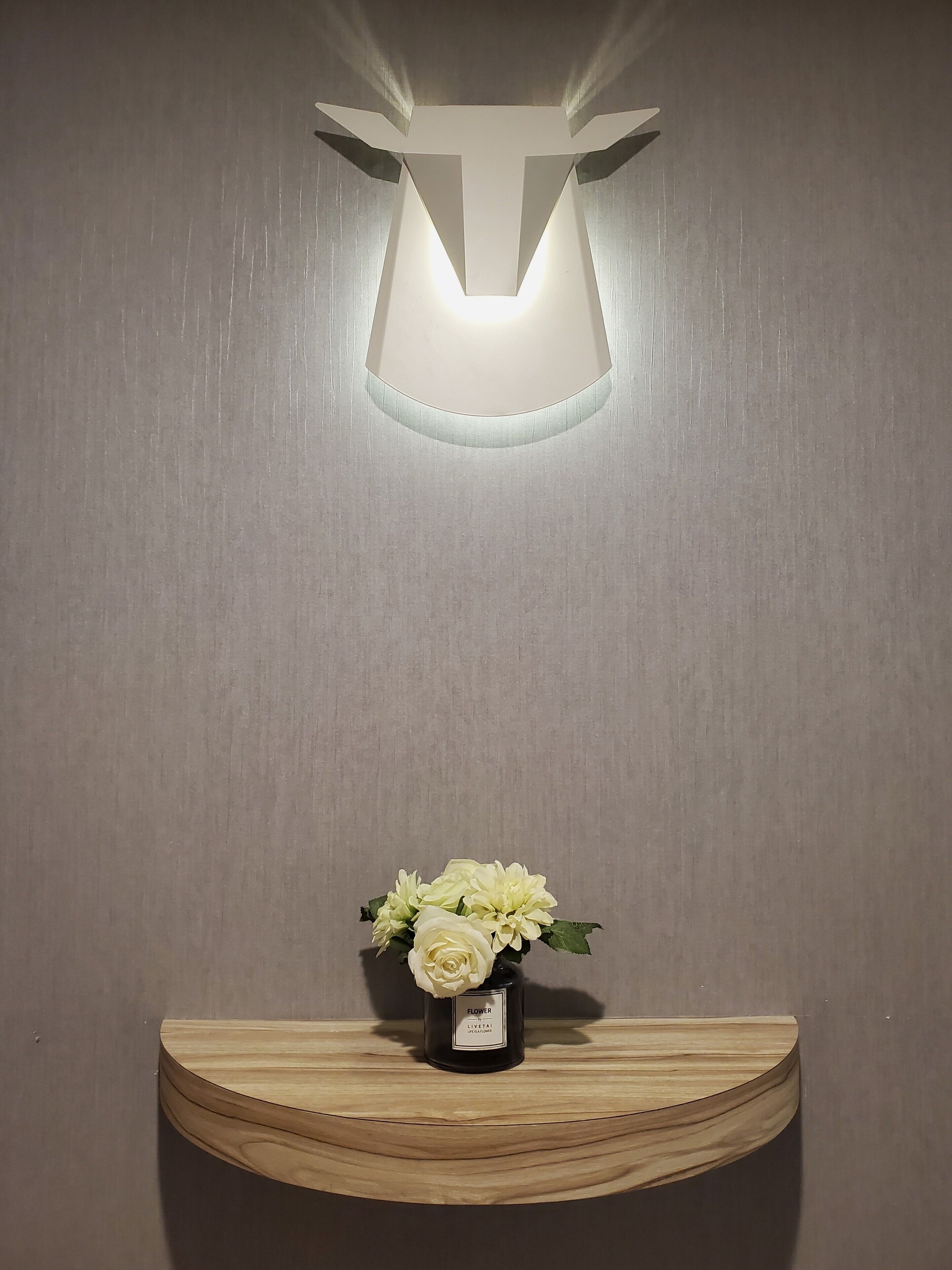 A wall-mounted light fixture with a geometric design and a wooden shelf below it holding a vase with white flowers.