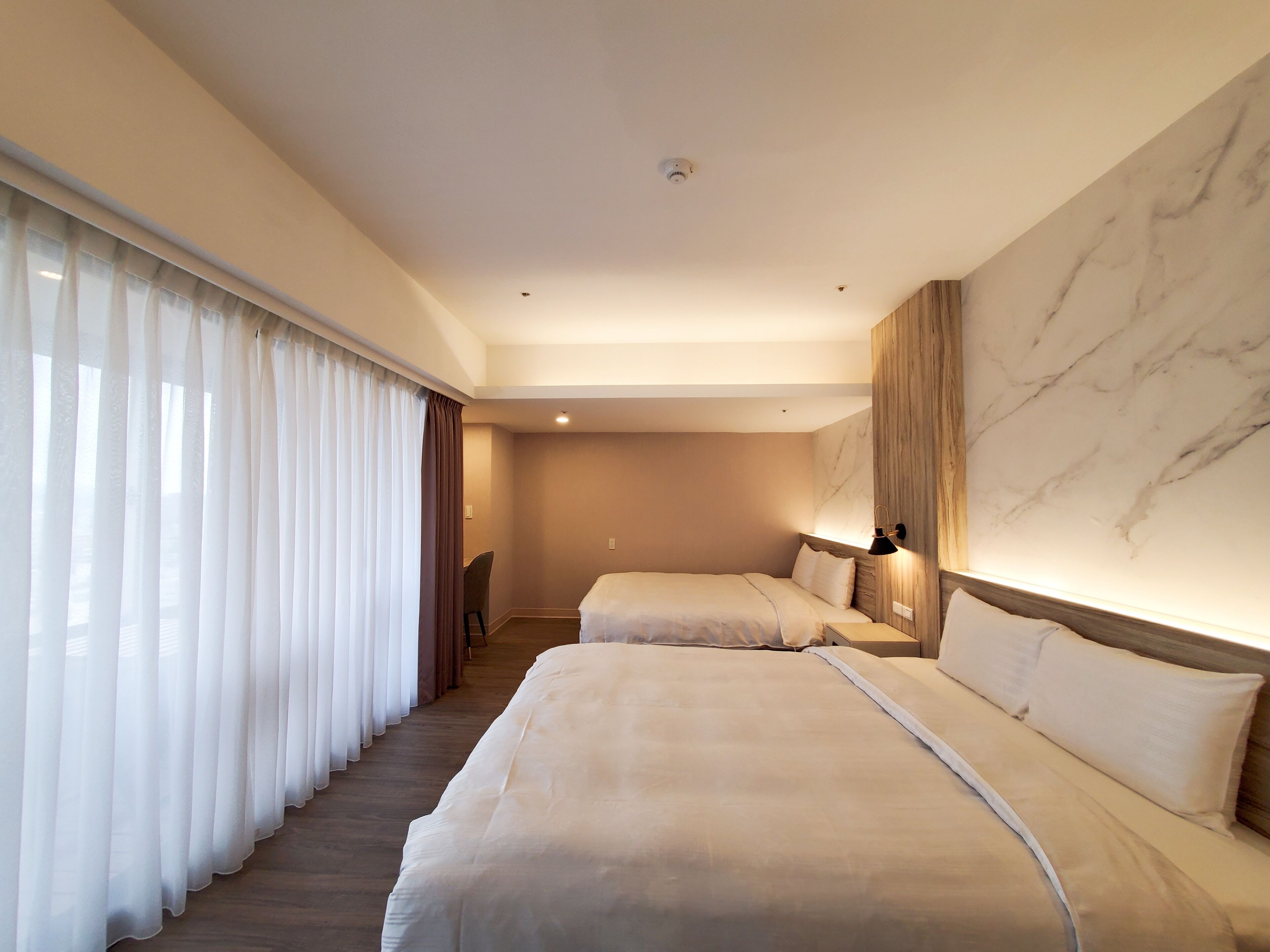A hotel room with two beds, a large window with sheer curtains, a marble accent wall, and a desk with a chair.