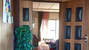 A wooden interior with a large potted plant, a framed picture, and a door leading to a living area with a sofa and a table.