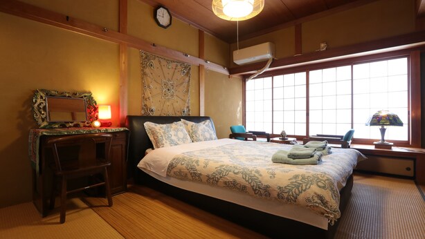 A traditional Japanese-style bedroom with tatami flooring, a large bed with patterned bedding, a desk with a lamp, and a view through large windows.
