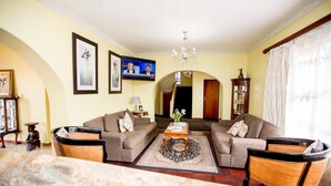 A living room with two sofas, a coffee table, and a TV. There are framed pictures on the wall, a chandelier, and a window with curtains. - Oria Guesthouse (Cape Town)