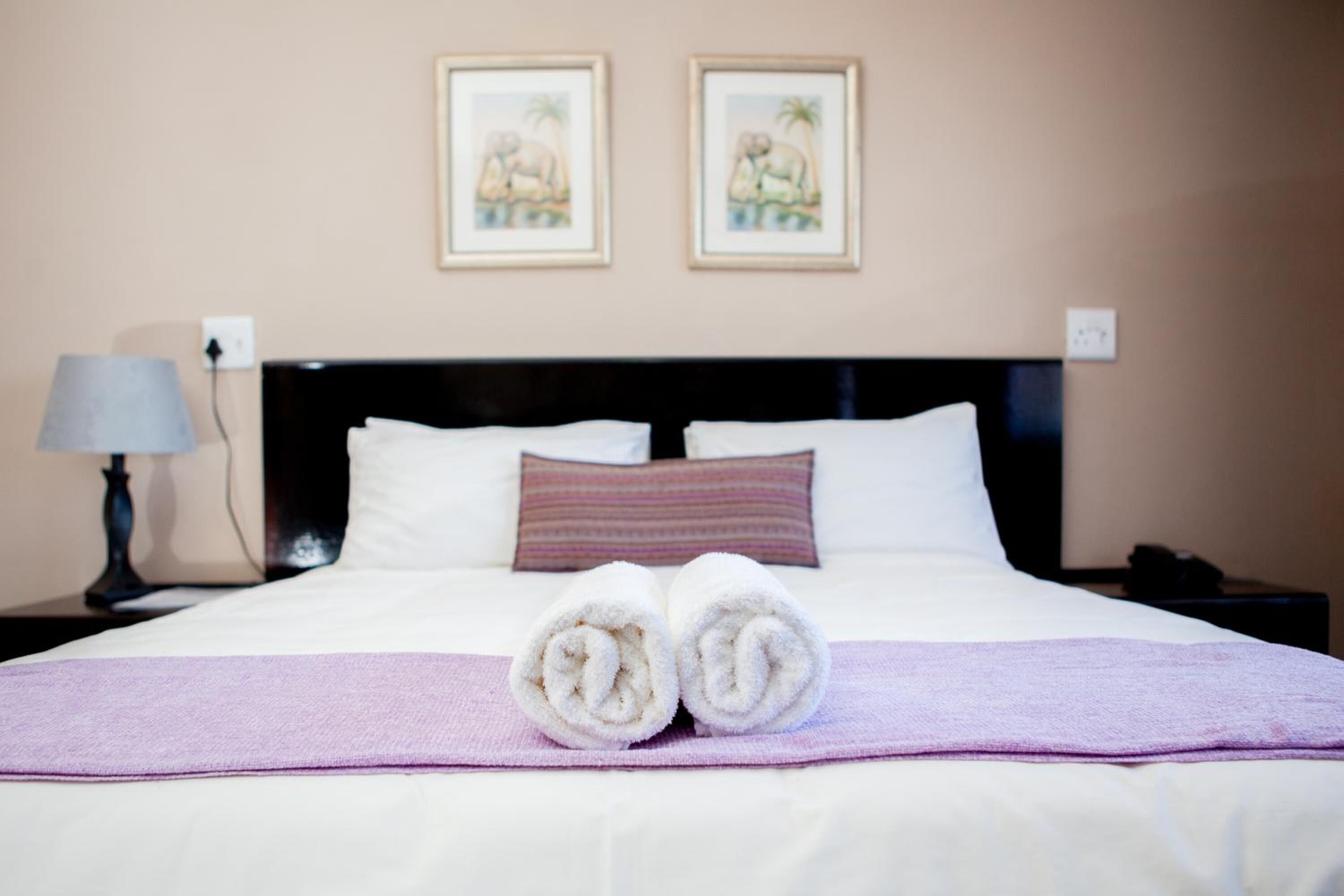 A neatly made bed with rolled towels, a purple throw blanket, and two framed pictures on the wall.