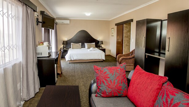 A hotel room with a bed, a sofa with red and grey pillows, a TV, a desk with a computer, and a wardrobe. - Oria Guesthouse (Cape Town)