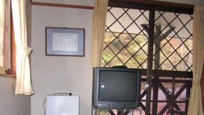 Una habitación con suelo de tatami, una mesa de madera, un televisor sobre un mueble, un calentador blanco y una ventana con vistas a árboles.