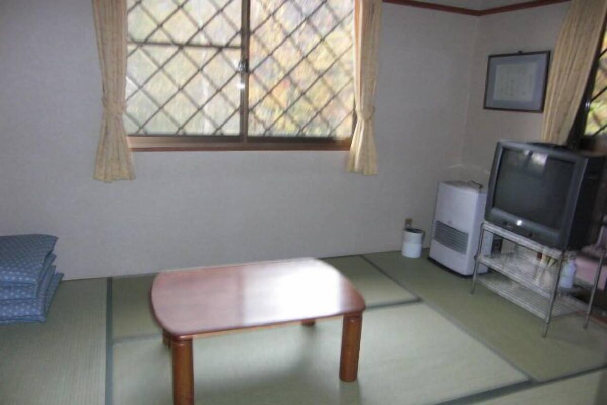 A room with tatami flooring, a low wooden table, a television on a stand, and a window with a curtain.