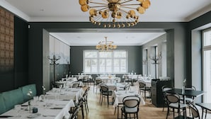 A dining area with multiple tables and chairs, a chandelier, and large windows allowing natural light. - Villa Santa Hotel (Cesis)