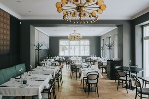 Un comedor con varias mesas y sillas, un candelabro y ventanas grandes que dejan entrar luz natural.