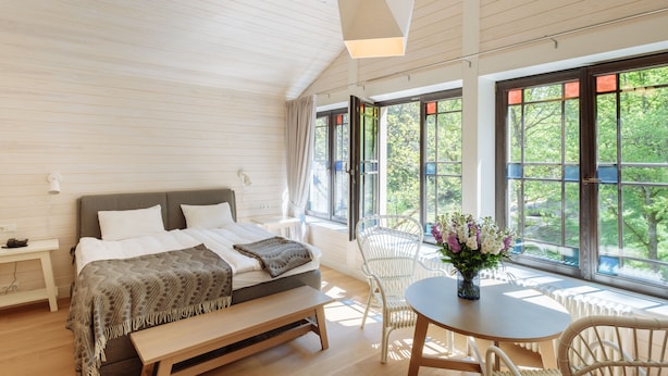 A bedroom with a bed, bedside table, and a view of greenery through large windows. - Villa Santa Hotel (Cesis)