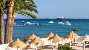 A coastal scene with palm trees, a beach with thatched umbrellas, and a marina with various boats. - Hotel Simbad (Ibiza Town)