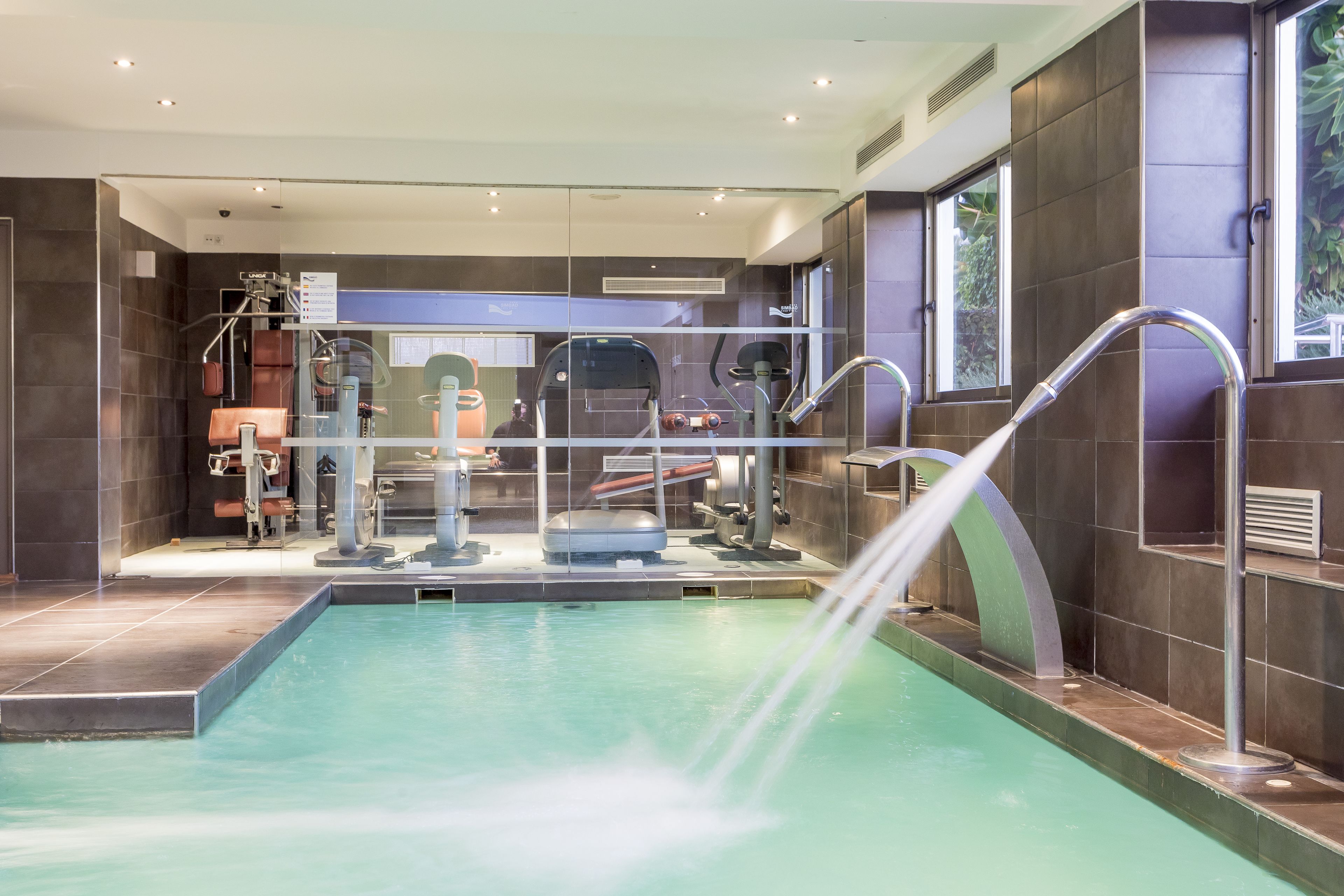 A modern indoor pool area with a water feature, exercise equipment, and a view of the outside.