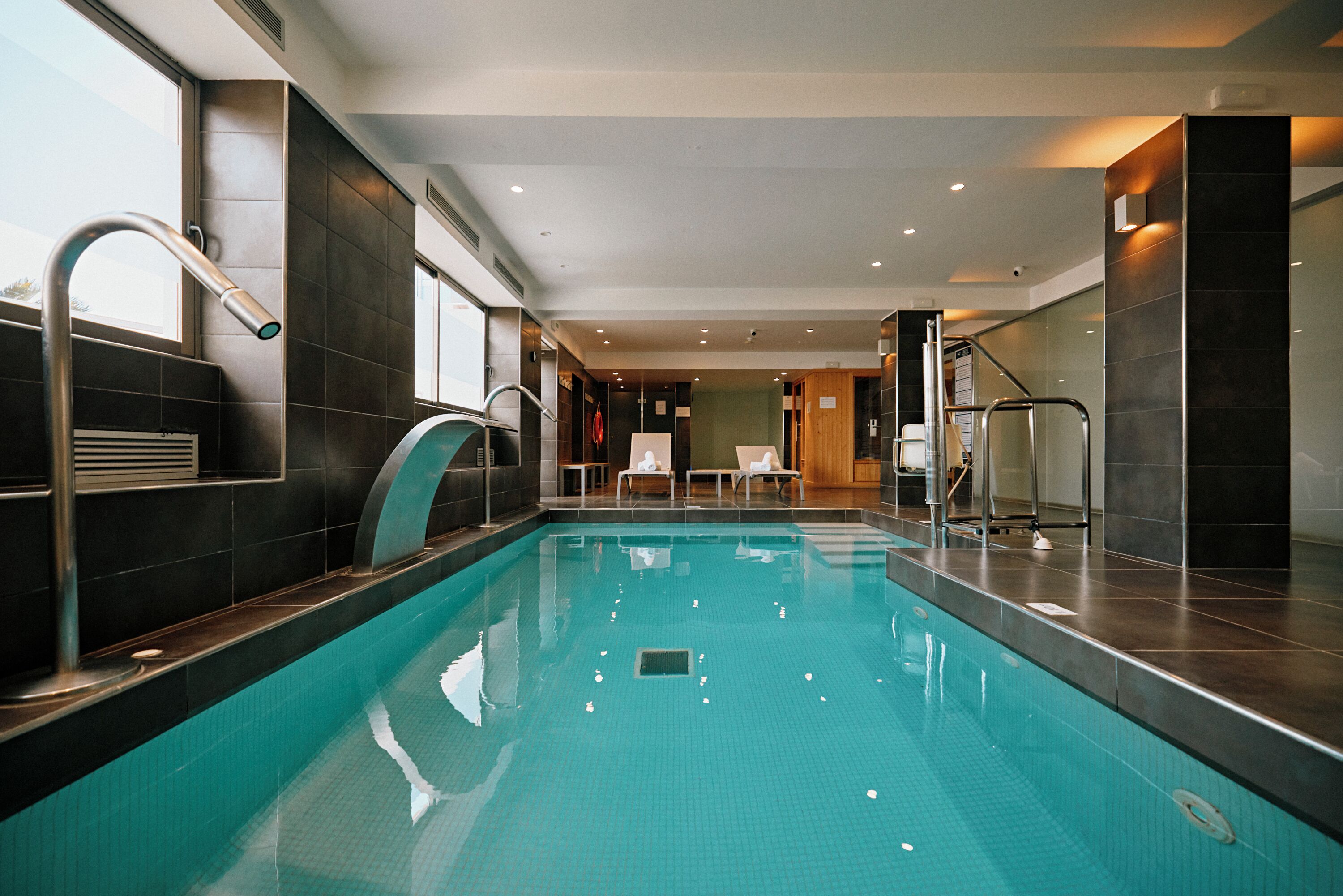 A modern indoor swimming pool with clear water, a metal ladder, and a seating area with chairs.