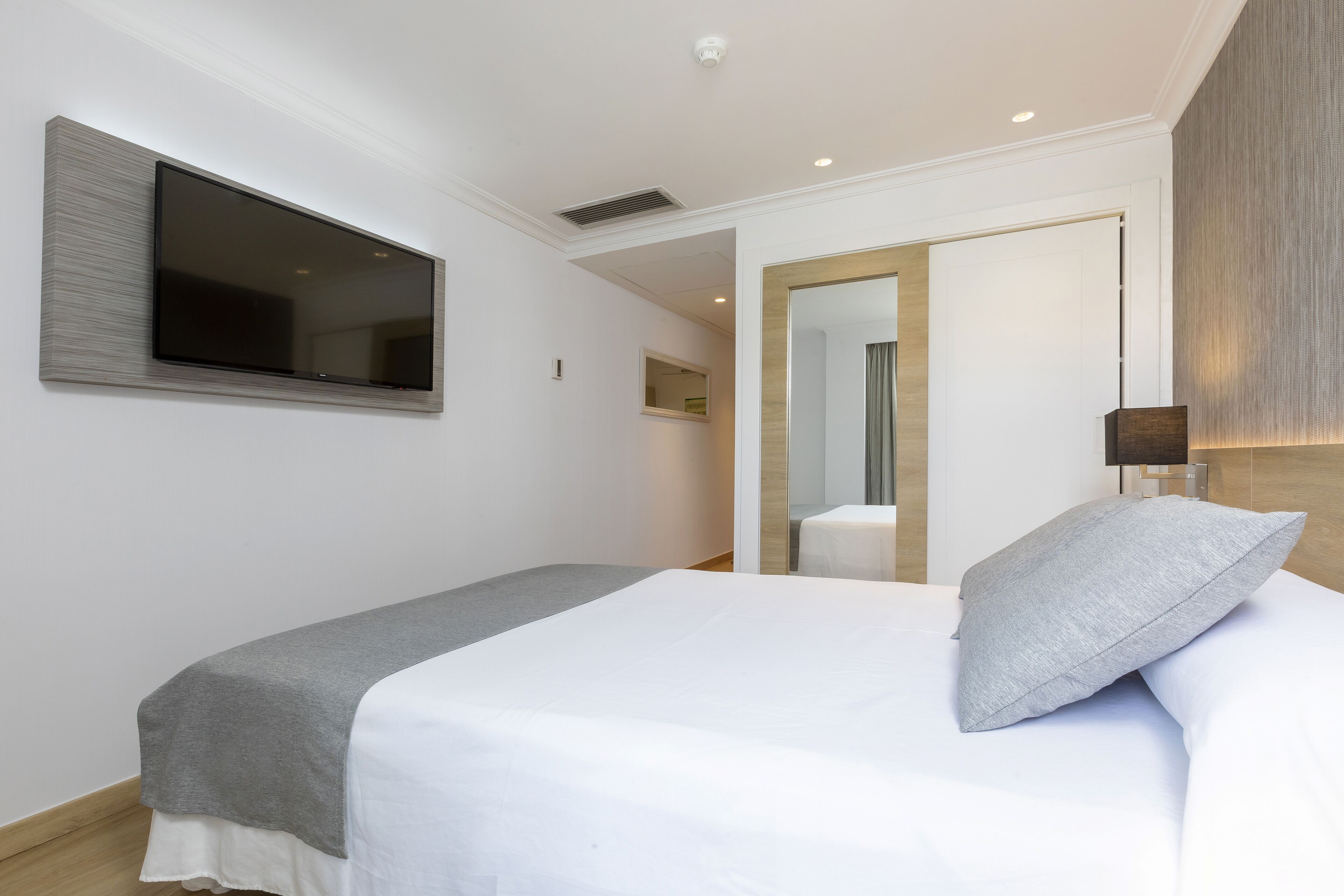 A modern hotel room with a large flat-screen TV mounted on the wall, a neatly made bed with a gray blanket, and a wooden headboard.