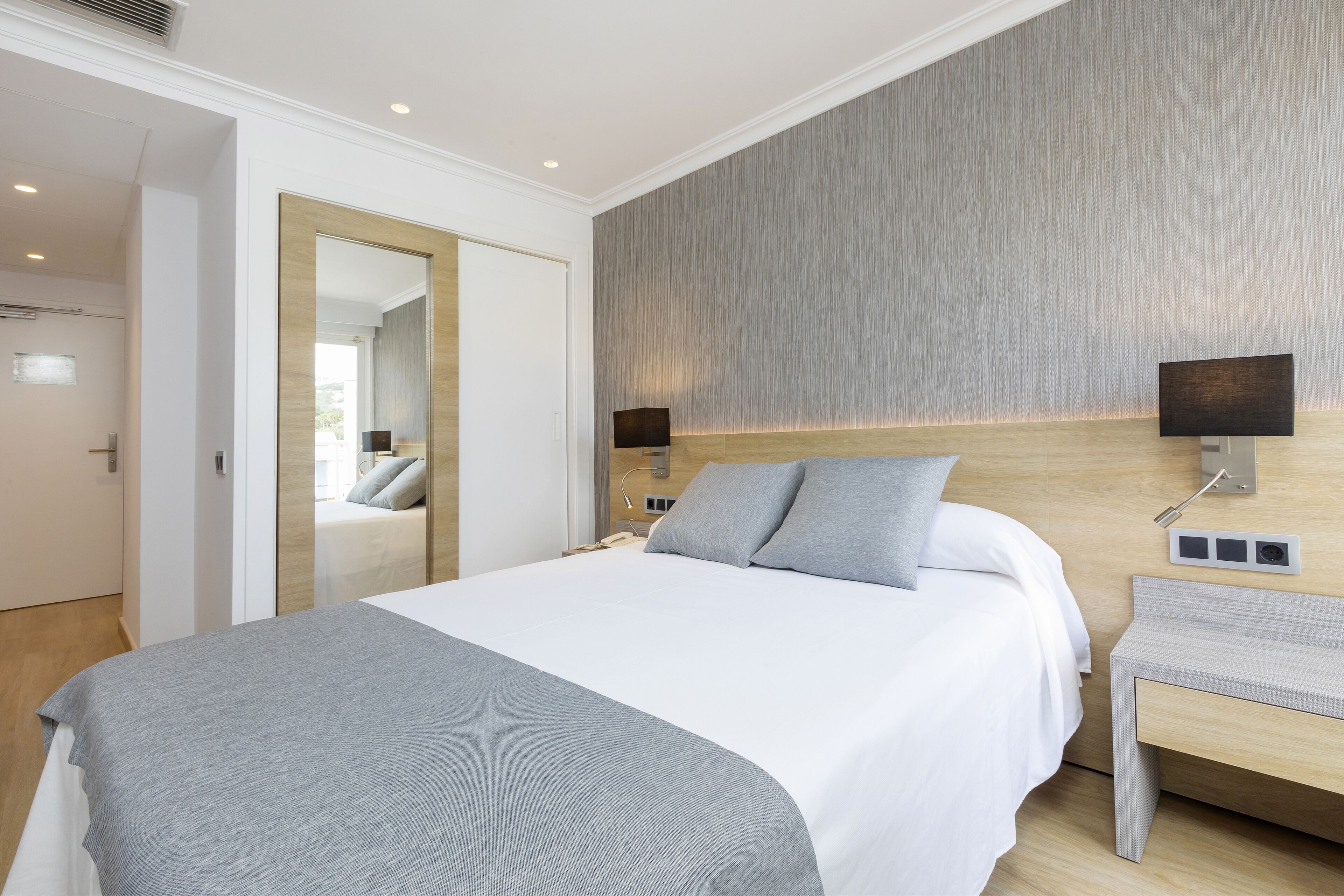 A modern hotel room with a large bed, a bedside table, a mirror, and a wall-mounted lamp.