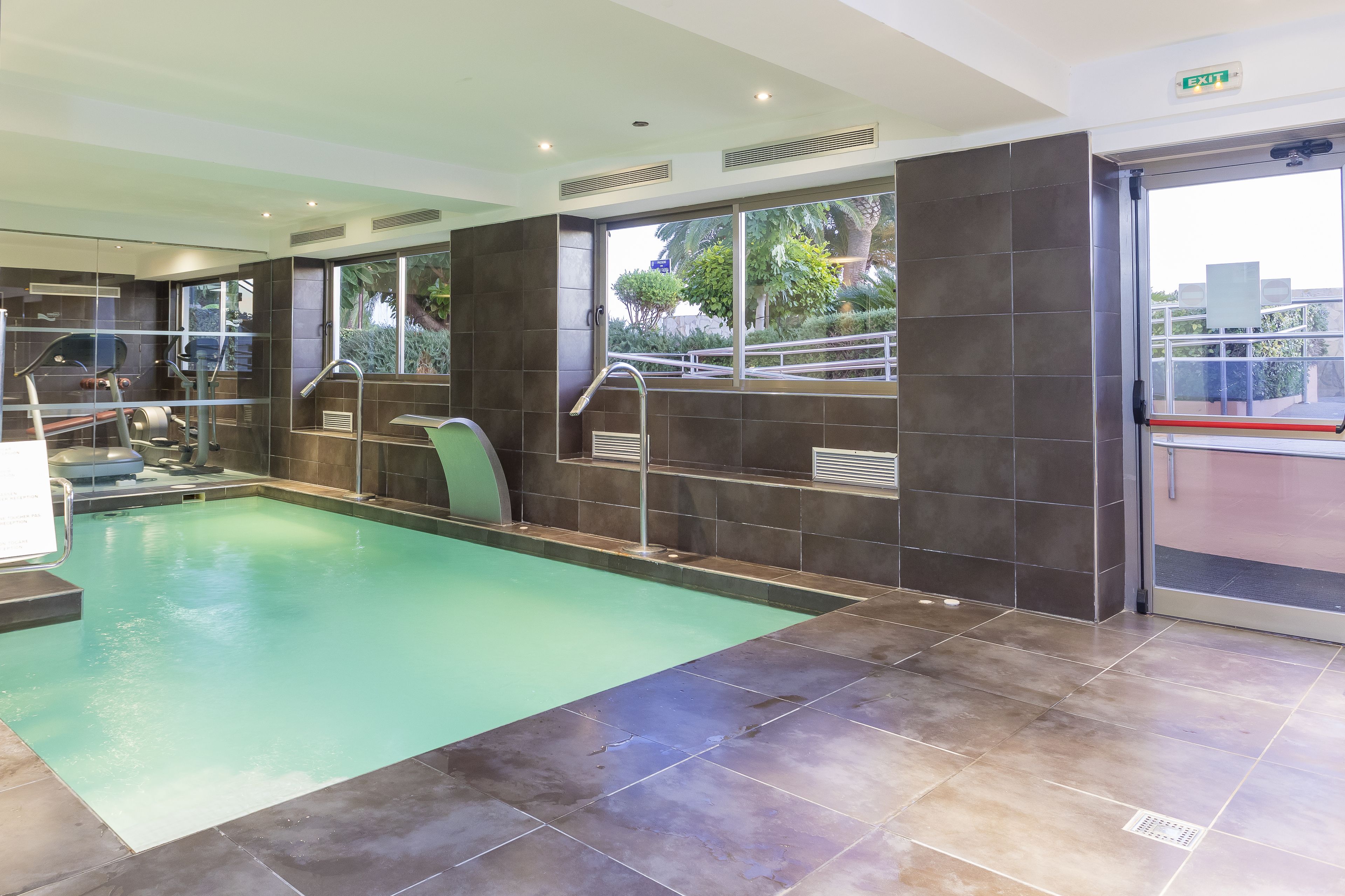A swimming pool with a glass wall, exercise equipment, and a view of greenery outside.