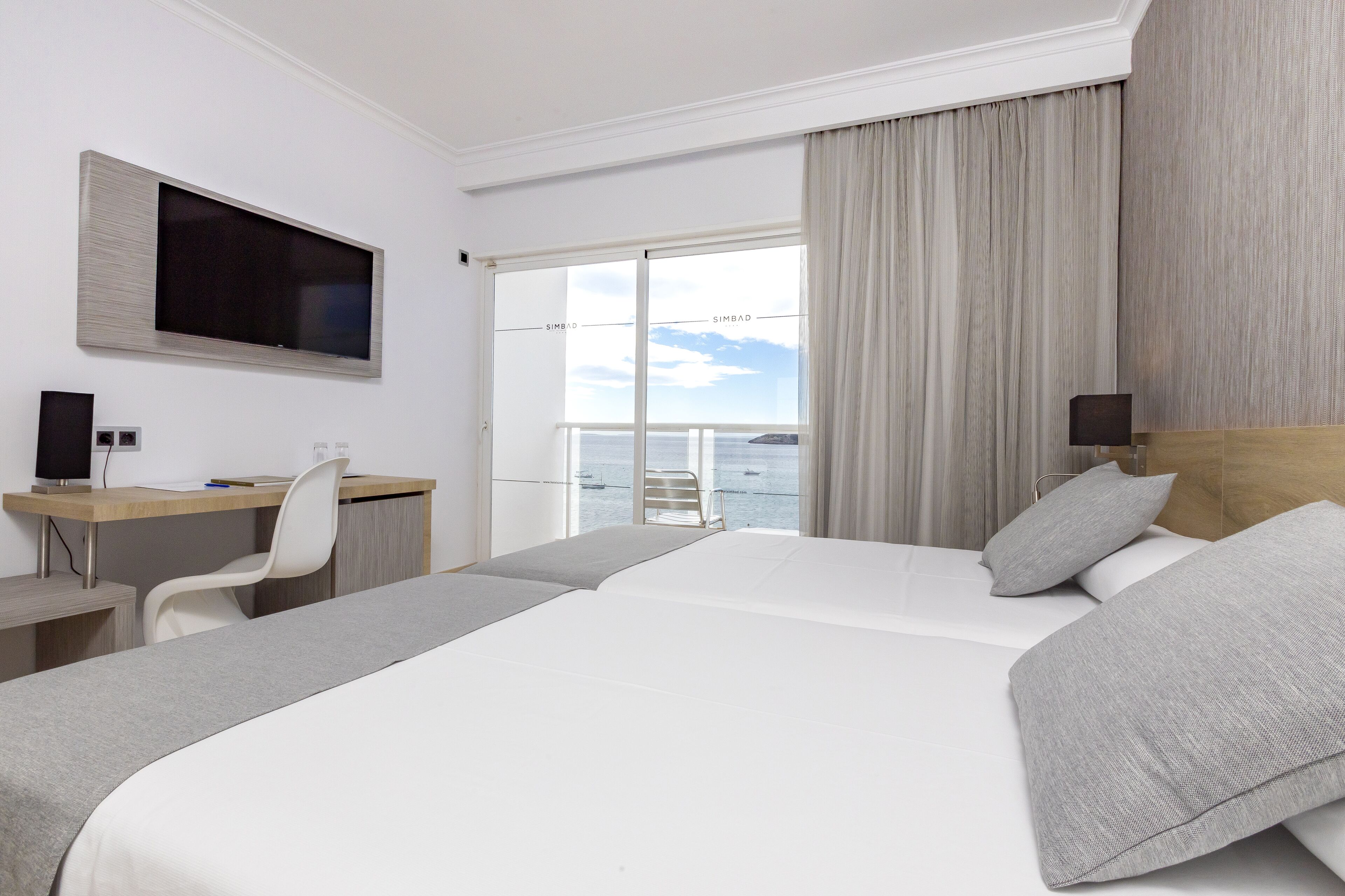 A hotel room with a large bed, a desk, a chair, a TV, and a view of the sea through a sliding door.