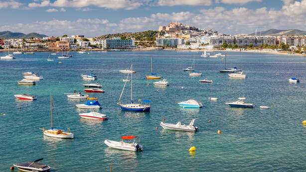 A coastal town with numerous boats docked in the marina, a prominent hilltop building, and a clear blue sky. - Hotel Simbad (Ibiza Town)