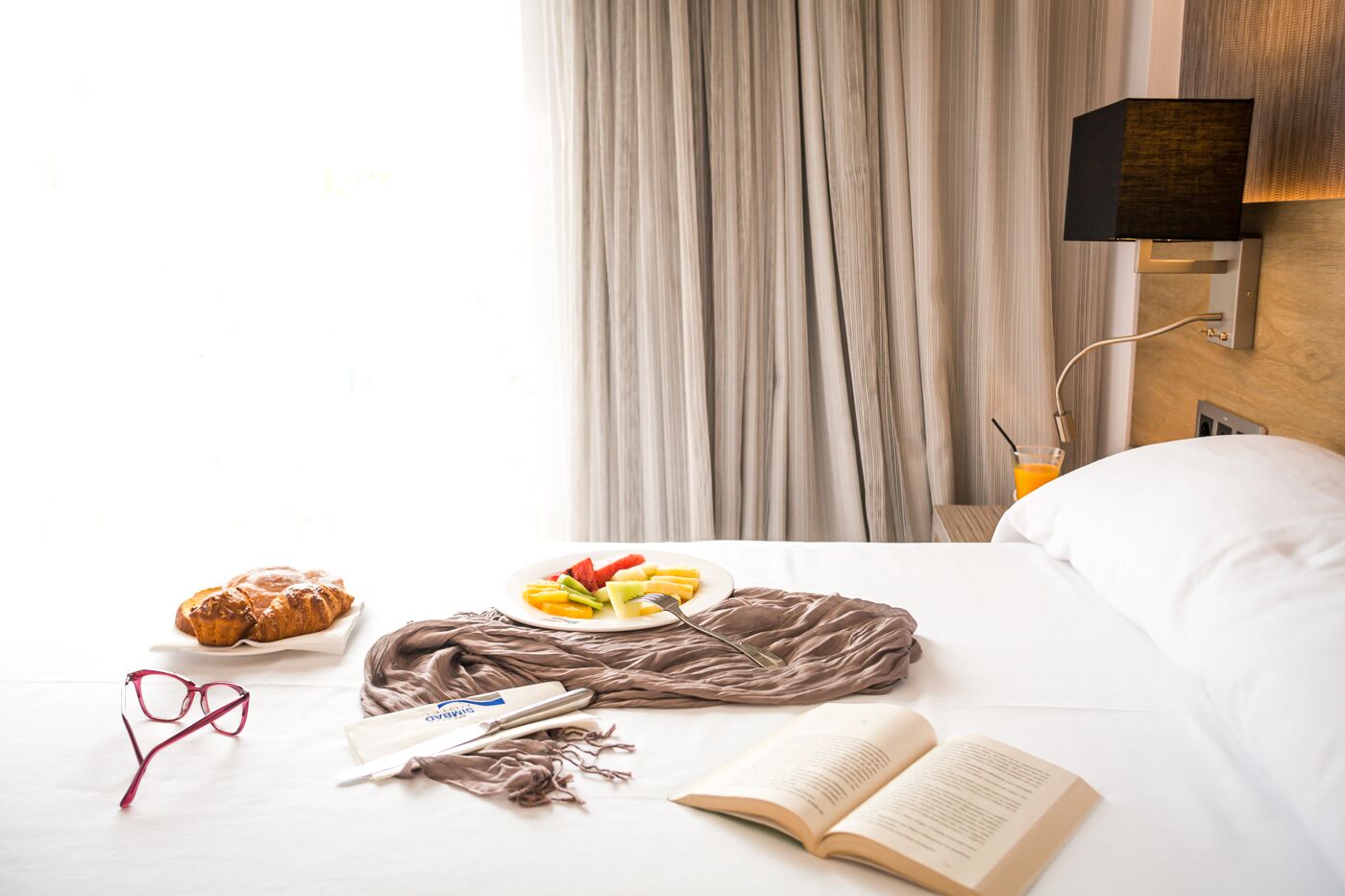 Una cama bien hecha con un almohadón, una mesita de noche con una lámpara, un plato de fruta, un croissant, un libro y un par de gafas.