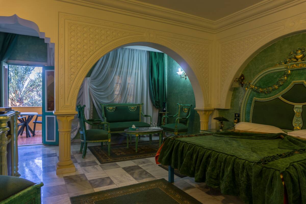 A room with a bed, two chairs, a small table, and an arched doorway leading to a balcony.