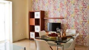 A modern dining area with a glass table, white chairs, and a television. A wooden bookshelf and a small shelf with decorative items are also visible. - Apartamento Ulises Suite 1 (Málaga)