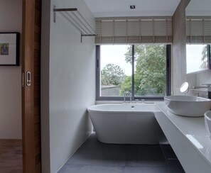 A modern bathroom with a freestanding bathtub, a large window with blinds, and a sink with a mirror.