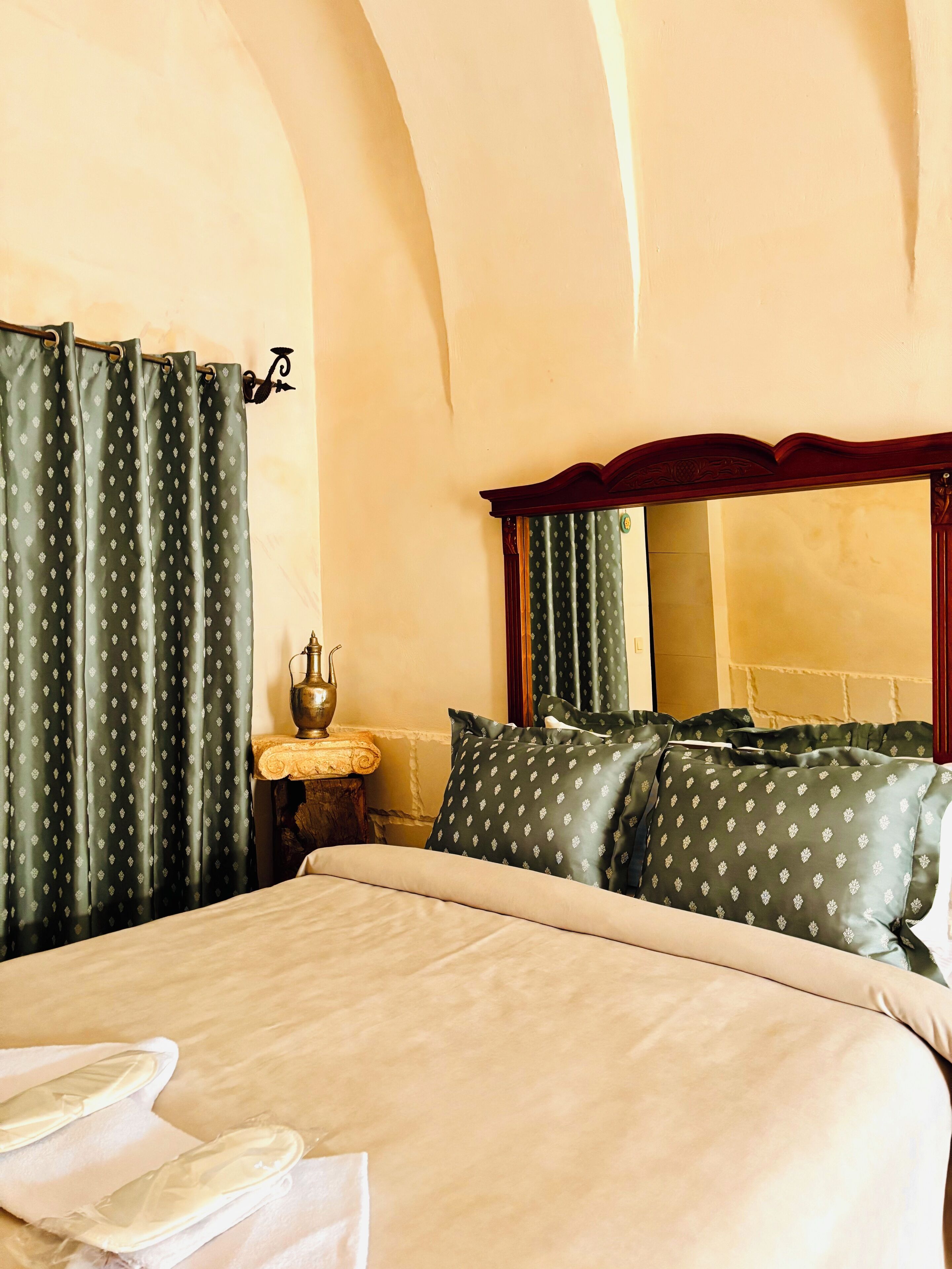 A hotel room with a large bed, green patterned curtains, a wooden headboard, and a small bedside table with a lamp.