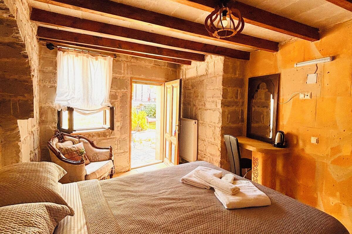 Un dormitorio rústico con vigas de madera a la vista, una cama grande con una manta texturizada, una silla y una mesita con una tetera.