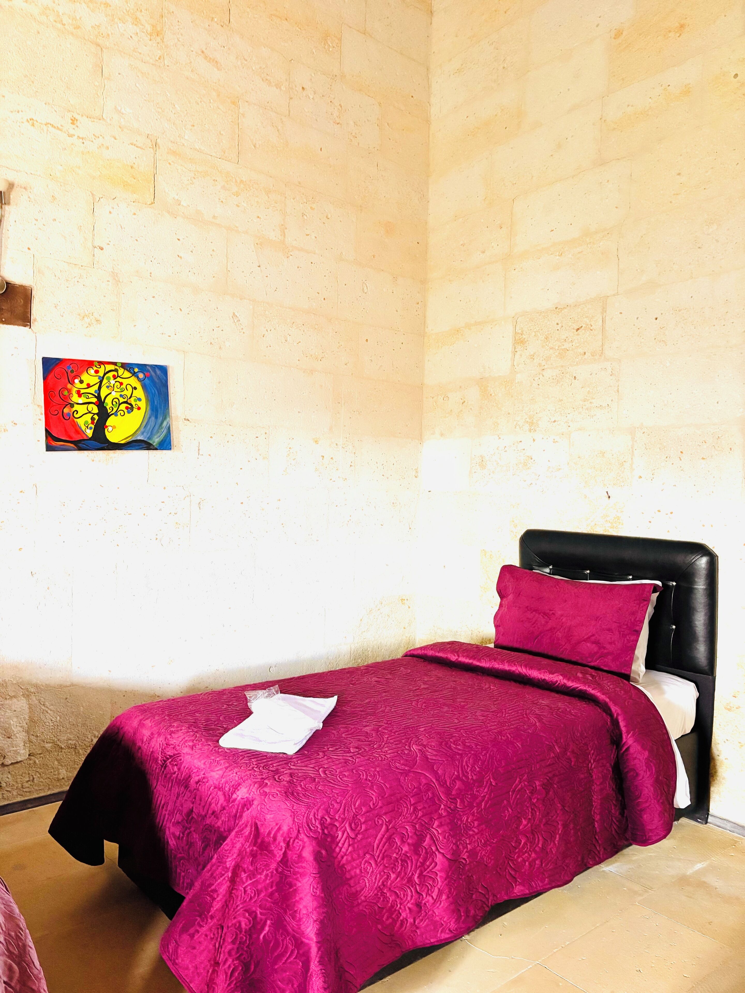 Ein kleines, ordentlich bezogenes Bett mit einem schwarzen Kopfteil und einer leuchtend roten Decke. Auf der Wand ist ein farbenfrohes Baumgemälde zu sehen.