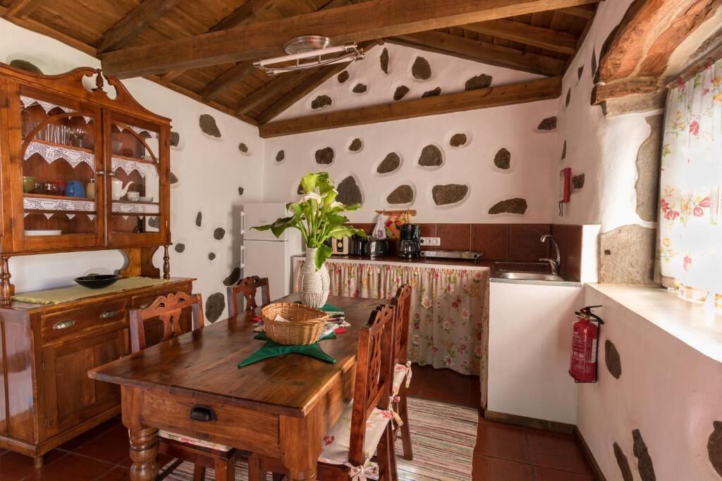 A rustic kitchen with a wooden dining table, chairs, a vase with a plant, and a stone wall.