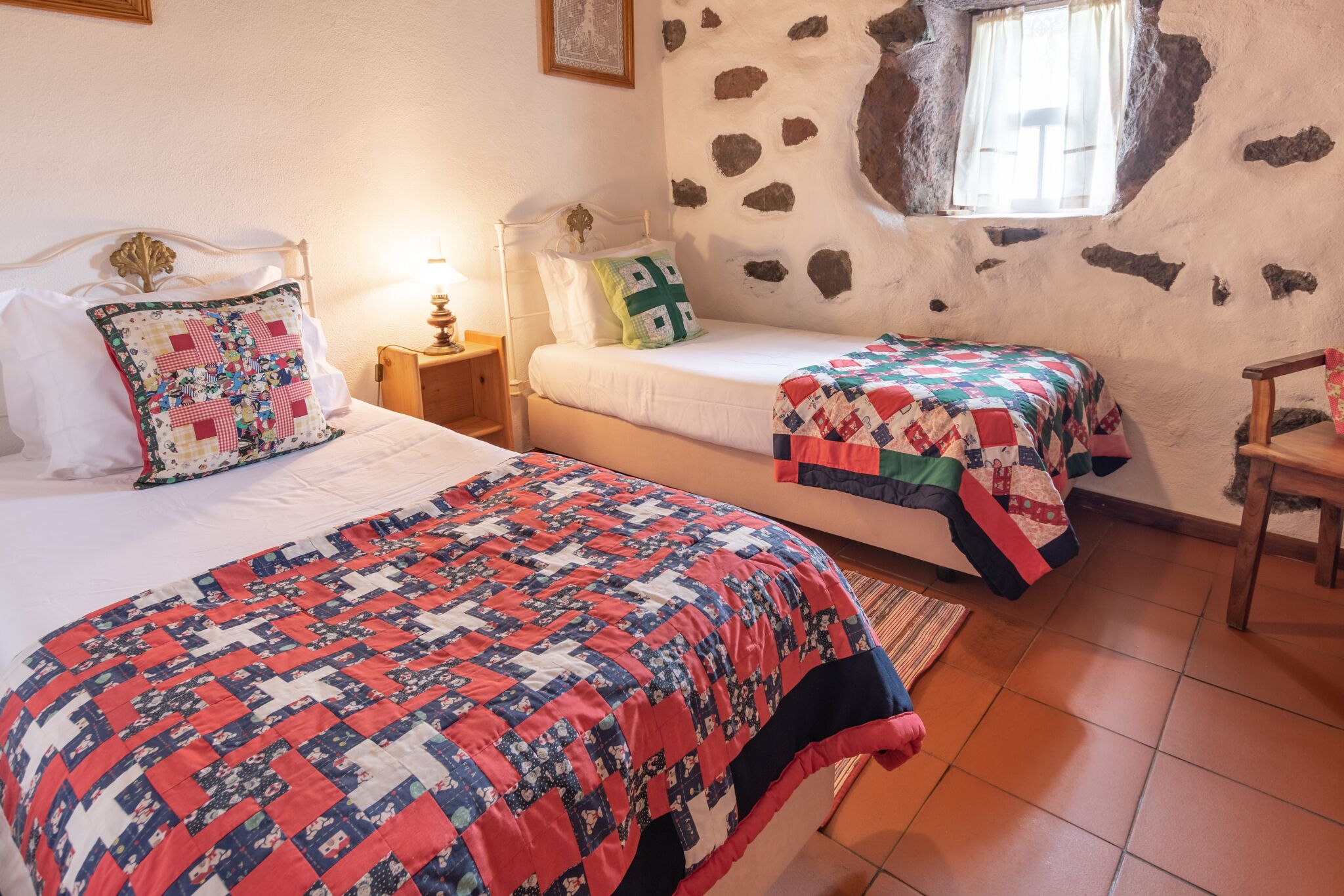 A bedroom with two beds, a stone wall, a window, and a wooden chair.